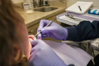 Dentist's gloved hands carefully performing root-planing on a patient's lower teeth in a warmly lit, authentic dental office, shot from the patient's perspective.