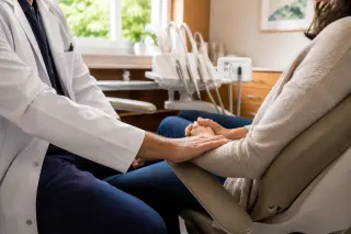 Dentist's hand resting on patient's forearm in a warm, softly lit dental office, both displaying relaxed, natural postures before a procedure