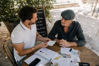 Osteopath and village shopkeeper sharing an authentic, focused exchange at an outdoor Provençal café table, with local flyers and notes visible, under soft afternoon sunlight.