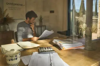 Sunlit Provençal osteopathy office with paperwork and osteopath reflecting on budget, natural and candid ambiance.