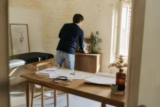 Young osteopath preparing a simple, sunlit Provençal office, arranging furniture and documents in a realistic, hopeful atmosphere.