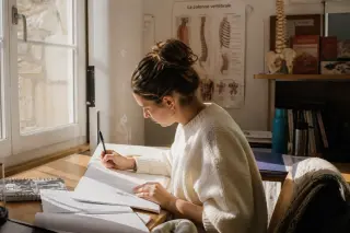 Osteopathy student studying handwritten notes by a window in a sunlit classroom with anatomical models in the background