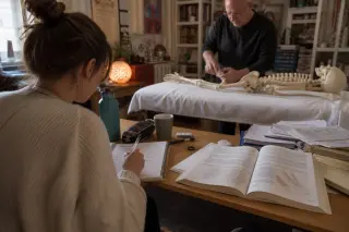 Osteopathy student intently writing notes while observing a practitioner's demonstration on a mannequin hand in a softly lit, authentic classroom with anatomical models and textbooks.
