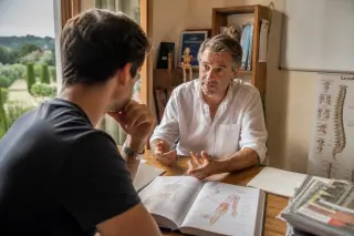 Osteopathy student and practitioner in a sunlit clinic discuss case studies at a wooden desk, with study materials and Mediterranean greenery visible through the window.
