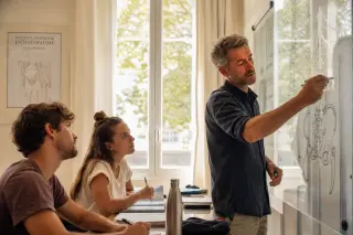 Osteopath instructor drawing on transparent board while attentive students take notes in a Provence seminar room with natural light