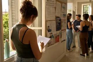 Osteopathy student in a sunlit corridor writing notes, other students chatting near a bulletin board, Provençal atmosphere.