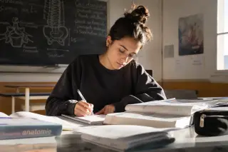 Focused student taking notes in a sunlit classroom with anatomy books and a chalkboard in the background, natural setting